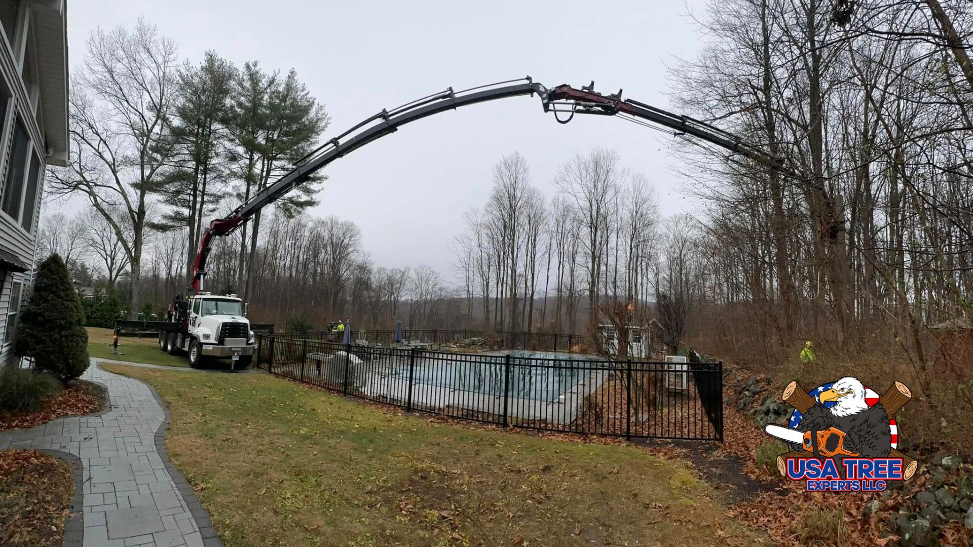 Copy of emergency (1920 x 1080 px) A large crane truck from a tree service company extends its hydraulic arm over a residential pool area to remove tree limbs from a wooded area in the backyard. A worker in high-visibility gear supervises the operation, demonstrating safe and precise tree removal near a home.
